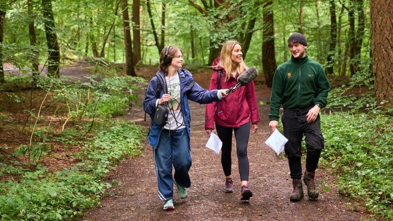 Ranger walking along a path in the woods together with a sound recorder holding a microphone, and an interviewer